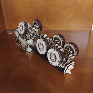 May include: A wooden model of a truck chassis with six wheels. The model is made of laser-cut wood and is assembled with small wooden pieces. The chassis is brown and the wheels are black.