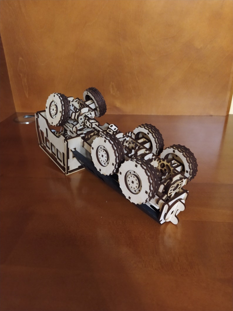 May include: A wooden model of a truck chassis with six wheels. The model is made of laser-cut wood and is assembled with small wooden pieces. The chassis is brown and the wheels are black.