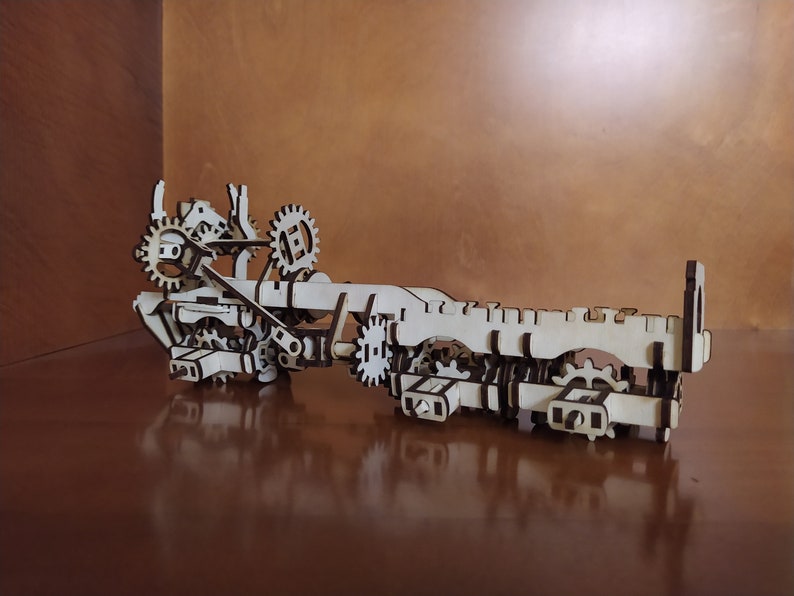 May include: A wooden model of a complex gear system with multiple interlocking gears and axles. The model is made of light brown wood and has a natural finish.