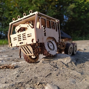 May include: A wooden model of a large truck with a detailed cab and a flatbed trailer. The truck is painted in a natural wood color and has black tires. The truck is sitting on a dirt road.