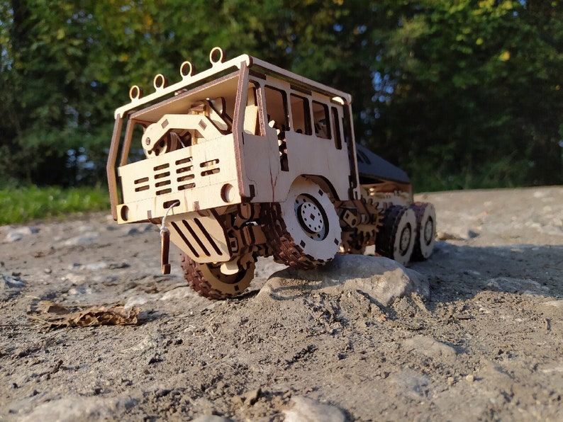 May include: A wooden model of a large truck with a detailed cab and a flatbed trailer. The truck is painted in a natural wood color and has black tires. The truck is sitting on a dirt road.