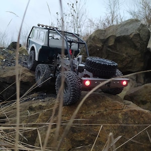 Puede incluir: Un camión de juguete blanco y verde con una jaula de seguridad negra y luces traseras rojas está conduciendo sobre rocas. El camión está en un terreno rocoso con hierba alta en primer plano.