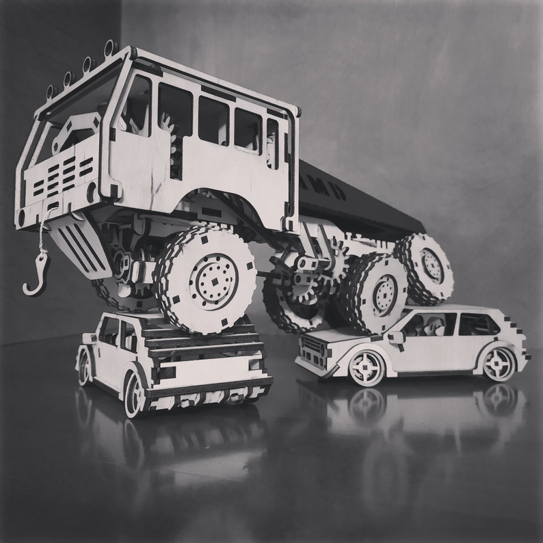 May include: A white wooden model of a large truck with a smaller car on top. The truck has a crane arm and a flatbed trailer. The car is a hatchback with a black interior.