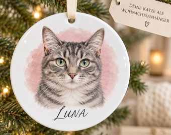 May include: A round white ceramic ornament featuring a watercolor-style portrait of a grey tabby cat with green eyes. The name Luna is printed in black script below the cat. A beige ribbon is attached to the top for hanging purposes.