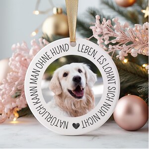 May include: White round ornament with a black border and the text "NATÜRLICH KANN MAN OHNE HUND LEBEN, ES LOHNT SICH NUR NICHT" in German. A photo of a golden retriever dog is in the center of the ornament.