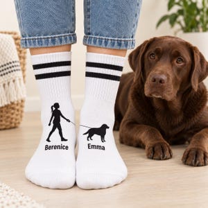 Könnte beinhalten: Weiße Crew-Socken mit zwei schwarzen horizontalen Streifen am oberen Rand. Die linke Socke zeigt eine schwarze Silhouette einer gehenden Person mit dem Namen Berenice, die rechte eine schwarze Hundesilhouette mit dem Namen Emma.