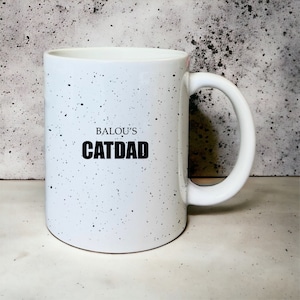 May include: White ceramic mug with black speckles and the text "BALOU'S CATDAD" printed on the front.
