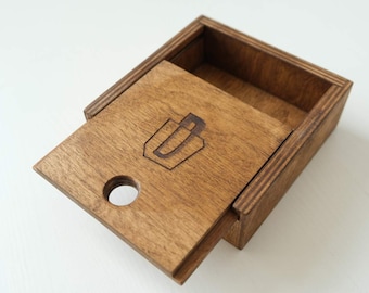 May include: A small wooden box with a sliding lid. The lid has a circular cutout and a laser-etched logo of a stylized letter "U". The box is stained a dark brown color.
