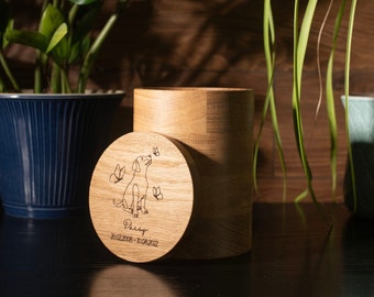 May include: A wooden pet urn with a personalized inscription that reads "Daisy 2020-2023". The urn features a laser-engraved image of a dog with butterflies. The urn is sitting on a dark wooden surface with two potted plants in the background.