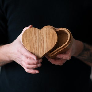 Puede incluir: Una caja de madera en forma de corazón con una tapa que se abre para revelar un interior hueco. La caja está hecha de madera marrón claro con un patrón de veta natural.