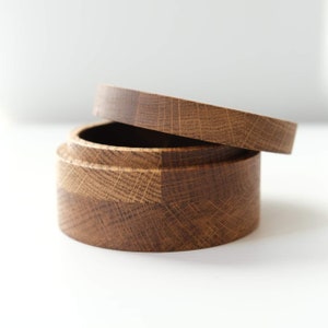 Op de afbeelding: Een ronde houten doos met een deksel, gemaakt van lichtbruin hout met een natuurlijke houtnerf. Het deksel is lichtjes open, waardoor de binnenkant van de doos zichtbaar is.
