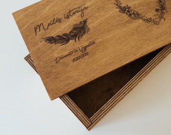 Puede incluir: Una caja de madera con tapa. La tapa está grabada con las palabras "meilės istorija" y "Dainavos ir Vygantas 20220729". La tapa también tiene un diseño de corazón floral. La caja está hecha de madera marrón claro.