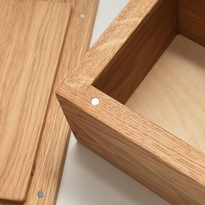 Puede incluir: Una caja de madera con una tapa con bisagras. La caja está hecha de madera marrón claro y tiene un acabado natural. La tapa está abierta y el interior de la caja es visible. El interior de la caja está forrado con un material marrón claro.