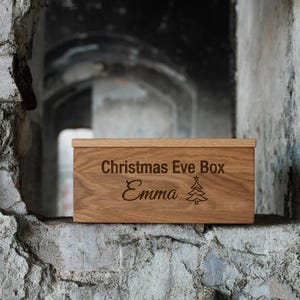 Könnte beinhalten: Eine rechteckige Holzbox mit hellbrauner Maserung. Auf der Vorderseite ist der Text "Christmas Eve Box" über dem Namen "Emma" und einem kleinen Tannenbaum-Symbol eingraviert. Der Deckel ist flach und passt zum Design der Box.