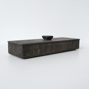 May include: A rectangular wooden box with a dark, weathered finish and a visible wood grain texture. The box features a flat, removable lid with a small, centered, square-shaped knob handle on top, providing a simple and functional storage design.