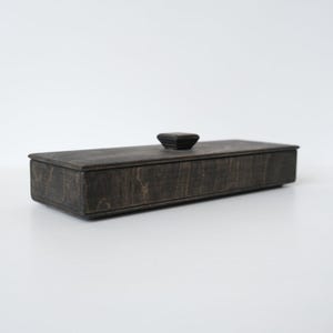 Peut inclure: Une boîte rectangulaire en bois avec une finition sombre et patinée, révélant le grain naturel du bois. Elle est dotée d'un couvercle plat amovible surmonté d'un petit bouton de préhension carré centré, offrant un design de rangement simple.