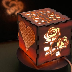 Puede incluir: Una lámpara de madera con forma de cubo con un corazón y rosas recortadas. La lámpara está encendida y proyecta un brillo cálido.
