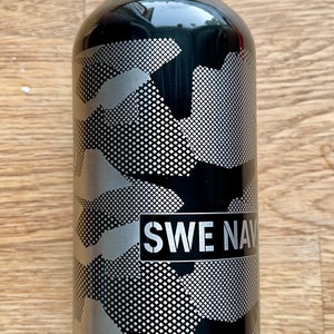 Könnte beinhalten: Schwarze Wasserflasche mit silbernem Tarnmuster und dem Text "SWE NAVY". Die Flasche ist zylindrisch und scheint aus Metall zu sein. Das Design weist ein gitterartiges Muster auf.