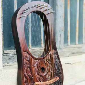 Può includere: Una lira in legno con intricate sculture di foglie e fiori. La lira ha 10 corde e un ponte in legno. La lira è marrone e ha una finitura naturale.