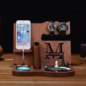 May include: A dark brown wooden docking station featuring a smartphone stand with a charging cable slot, two watch holders, and a wallet compartment. The front panel is engraved with a large letter M and the name Michael, alongside small chess pieces.