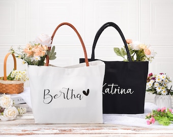 Op de afbeelding: Twee gepersonaliseerde tote bags, een witte met bruine handvatten en de naam "Bertha" met een hartje, en een zwarte met zwarte handvatten en de naam "Katrina".