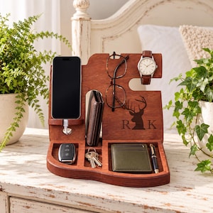 May include: A dark brown wooden docking station featuring a smartphone stand with a charging cable, a glasses holder, a watch hook, and compartments for a wallet, keys, and a pen. The wood is engraved with a deer silhouette and the letters R K.