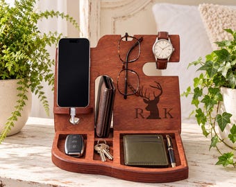 May include: A dark brown wooden docking station featuring a smartphone stand with a charging cable, a glasses holder, a watch hook, and compartments for a wallet, keys, and a pen. The wood is engraved with a deer silhouette and the letters R K.