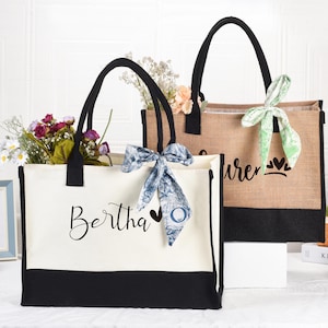Op de afbeelding: Twee gepersonaliseerde tote bags met zwarte handvatten en een zwarte onderrand. De tas aan de linkerkant heeft een witte achtergrond met de naam "Bertha" en een zwart hart. De tas aan de rechterkant heeft een bruine jute achtergrond met de naam "Lauren" en twee zwarte harten. Beide tassen hebben een groen-witte gepatenteerde sjaal die om de handvatten is gebonden.