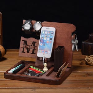 Könnte beinhalten: Ein dunkelbrauner Schreibtisch-Organizer aus Holz mit Telefon, Geldbörse, Schlüsseln und Uhren. Der Organizer hat ein Fach für kleine Gegenstände und einen Schlitz, damit das Telefon aufrecht stehen kann. Die Uhren werden auf einem Holzhalter ausgestellt.