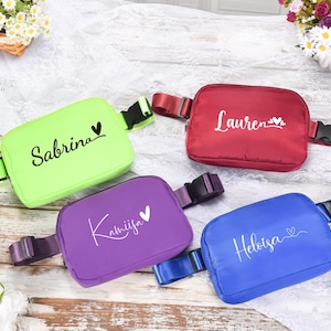 Könnte beinhalten: Vier bunte Bauchtaschen mit verstellbaren Riemen. Jede Tasche hat einen anderen Namen darauf gedruckt: Sabrina, Kamijfa, Helbisa und Lauren. Jeder Name wird von einem kleinen Herzen begleitet.
