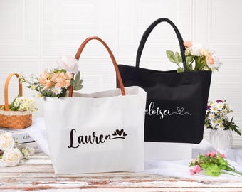 Op de afbeelding: Twee witte canvas tassen met bruine en zwarte handvatten. De tassen hebben zwarte letters met de namen "Lauren" en "Eloisa" en kleine hartjes.