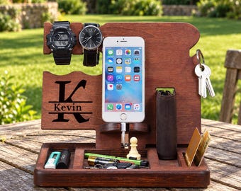 May include: A dark brown wooden docking station featuring a large letter K and the name Kevin engraved on the front. It includes slots for a smartphone, two wristwatches, a wallet, keys, and small accessories like a pen and cards.