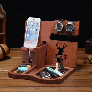 May include: A brown wooden docking station featuring a smartphone stand, a watch holder for two timepieces, and a wallet slot. The front panel displays a black silhouette of a deer head above the name David. The base includes compartments for small items like keys, a battery, and a miniature Eiffel Tower replica.