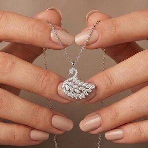 May include: A silver-coloured necklace featuring a swan-shaped pendant encrusted with numerous small, clear, marquise-cut stones. The pendant is suspended from a delicate, thin silver-coloured chain that drapes downwards, showcasing the intricate design.