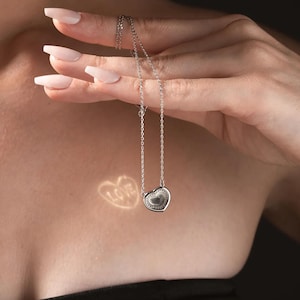 Puede incluir: Un collar de cadena de plata con un colgante en forma de corazón. El colgante tiene una superficie texturizada y una pequeña piedra transparente en el centro. El collar está siendo sostenido por una mano con uñas cuidadas.