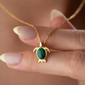 May include: A gold-colored necklace featuring a turtle pendant with a dark green stone inlay. The turtle pendant has a detailed design with flipper-like arms and legs. The necklace is held by a person.