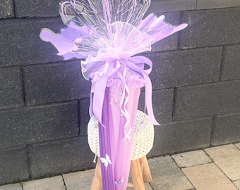 Könnte beinhalten: Eine lila-weiße Schultüte, verziert mit Schmetterlingsakzenten und einem großen, transparenten Schmetterlingsflügel-Design. Der Kegel ist mit einem lila Band gebunden und steht auf einem kleinen Holzschemel.