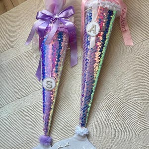 Könnte beinhalten: Zwei Schultüten im Meerjungfrauen-Design. Die Tüten sind mit schillernden Pailletten bedeckt und mit Bändern und Tüll verziert. Eine hat ein "S" und die andere ein "A" auf der Vorderseite. Die Basis jeder Tüte hat ein Meerjungfrauenschwanz-Design.