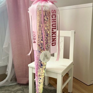 Könnte beinhalten: Eine rosa-goldene Schultüte im Meerjungfrauen-Design mit einer paillettenbesetzten Schwanzflosse und einem dekorativen Band. Der Kegel ist mit dem Text "SCHULKIND 2024" und dem funkelnden Buchstaben "A" verziert.