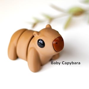 May include: A small, brown, hand-painted capybara figurine with black eyes and a red nose. The figurine is sitting on a white surface. The text "Baby Capybara" is written below the figurine.