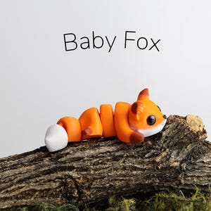 May include: A small orange and white fox figurine made of polymer clay, lying on a brown log. The figurine has a curled tail and large black eyes. The text "Baby Fox" is written above the figurine.