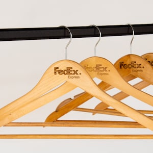 May include: A set of five wooden hangers with a brown finish. Each hanger has a silver hook and the FedEx Express logo is printed on the top of each hanger.
