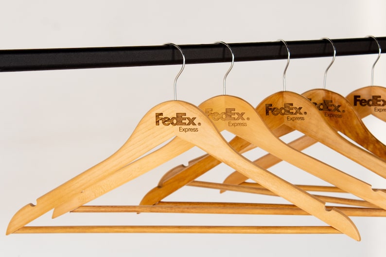 May include: A set of five wooden hangers with a brown finish. Each hanger has a silver hook and the FedEx Express logo is printed on the top of each hanger.
