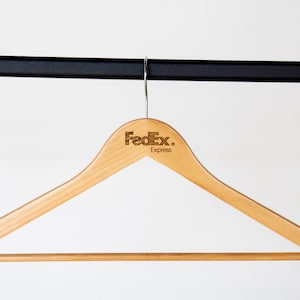 May include: A wooden clothes hanger with a brown finish. The hanger has a metal hook and a bar for hanging clothes. The hanger is branded with the FedEx Express logo.