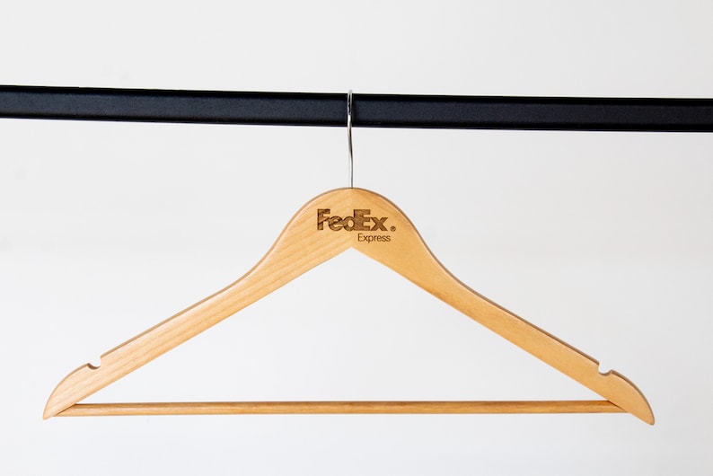 May include: A wooden clothes hanger with a brown finish. The hanger has a metal hook and a bar for hanging clothes. The hanger is branded with the FedEx Express logo.