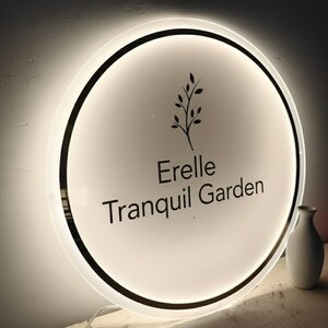 May include: A round, illuminated sign with the text "Erelle Tranquil Garden" in black. The sign features a black border and a stylized plant graphic. A small white vase is visible to the right.