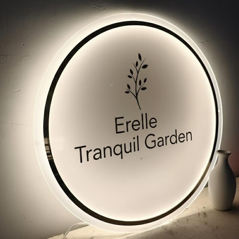 May include: A round, illuminated sign with the text "Erelle Tranquil Garden" in black. The sign features a black border and a stylized plant graphic. A small white vase is visible to the right.