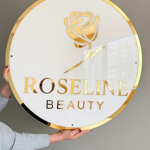 Puede incluir: Un letrero redondo con fondo blanco y borde dorado. El letrero presenta un gráfico de rosa dorada y las palabras "ROSELINE BEAUTY" en letras doradas. El letrero está sostenido por una persona.