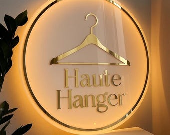 May include: A round, illuminated sign with a gold-colored border and a gold hanger graphic. The words "Haute Hanger" are in gold lettering below the hanger. The background is a soft, warm color.