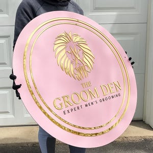 May include: Pink, oval sign with gold trim and a gold lion emblem. The sign reads "THE GROOM DEN EXPERT MEN'S GROOMING" in gold lettering. The sign is held up by a person.